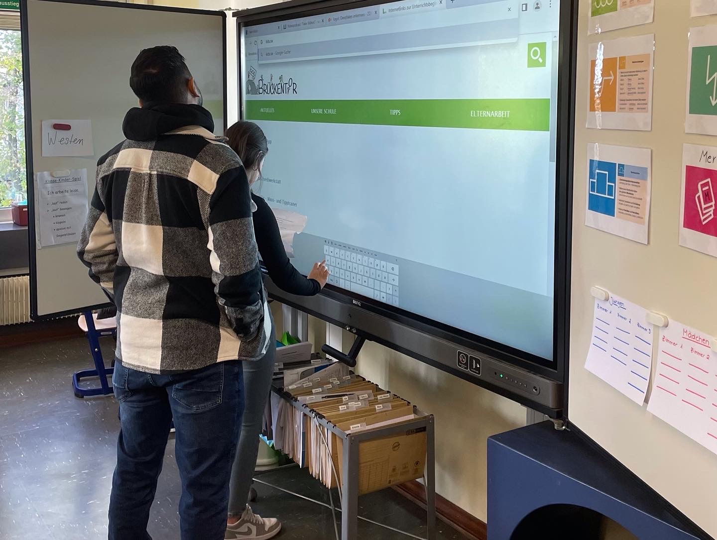 Bild von zwei Auszubildenden vor smart Screen und erklären Schülern im Klassenraum