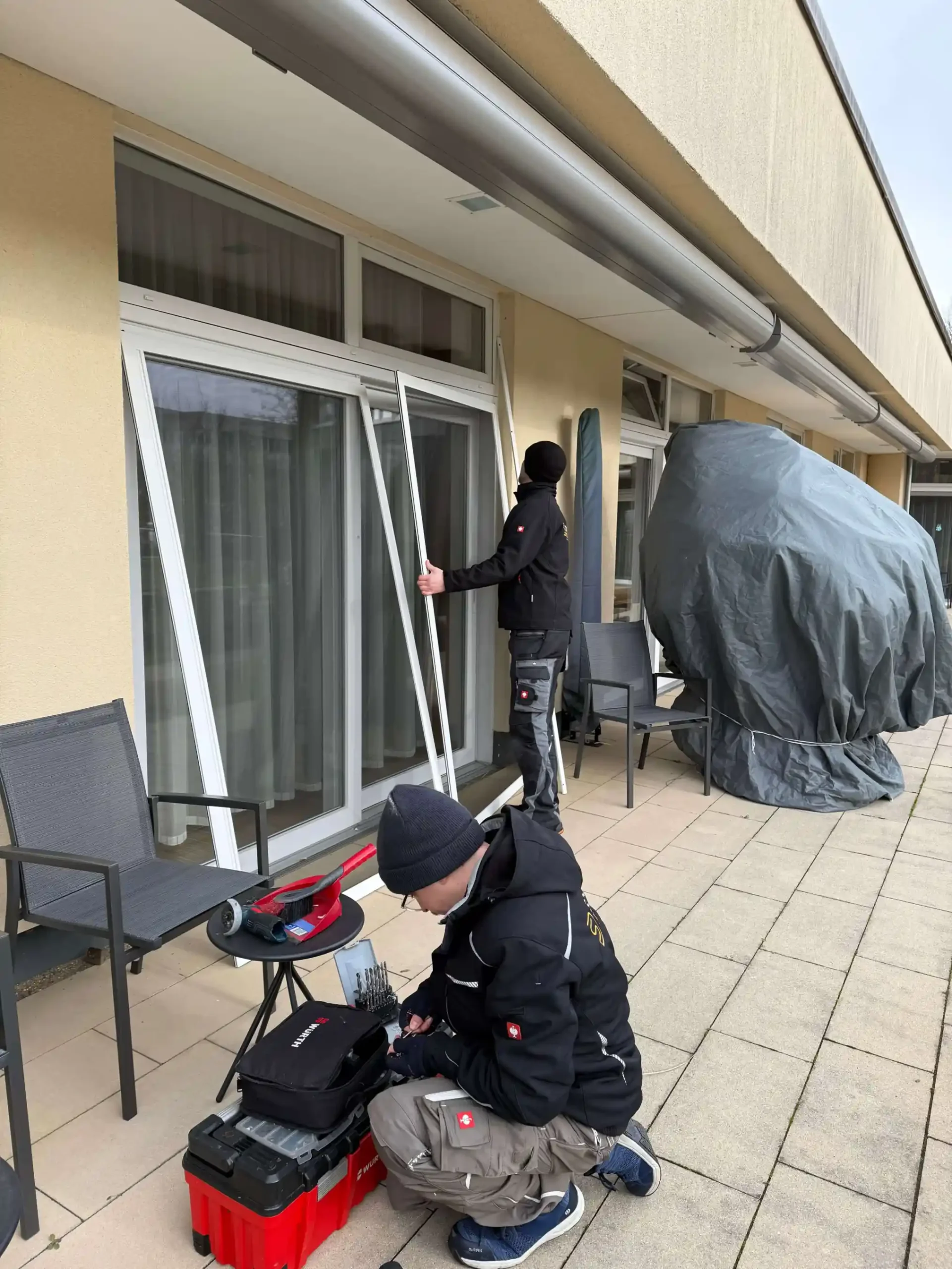 Zwei Monteure bauen eine weiße Insektenschutz-Schiebeanlage ein. Einer kniet an seinem Werkzeugkasten. Der andere bewegt das Insektenschutzgitter.