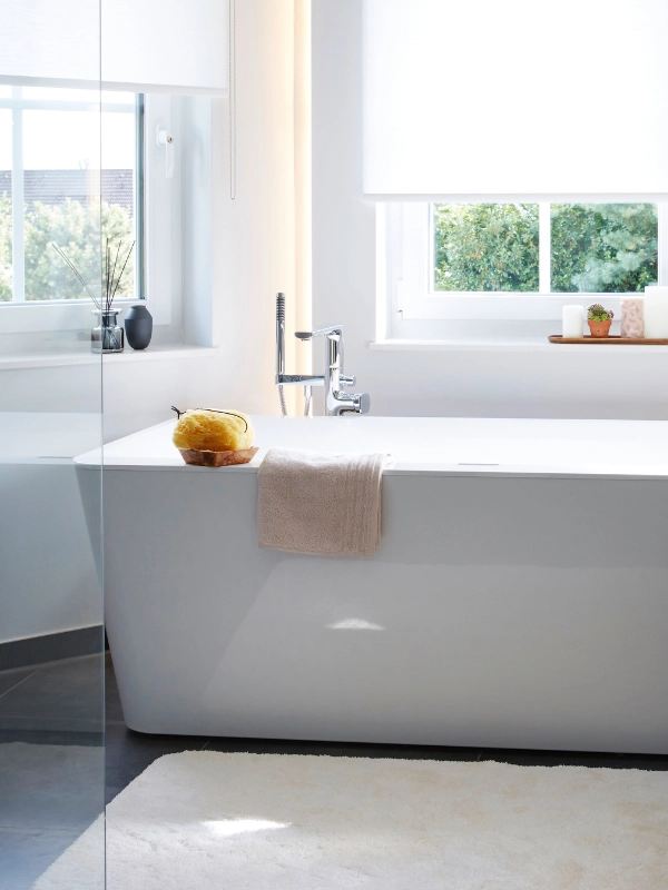 Zwei Badezimmerfenster sind mit weißen Rollos versehen. Die Rollos sind halb heruntergezogen. In der Mitte steht eine freistehende, weiße Badewanne. Links am Bildrand sieht man noch die Glaswand einer Dusche.