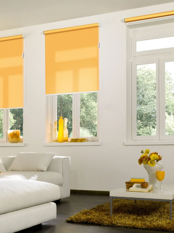 Ein weißes Wohnzimmer mit gelben Deko-Farbakzenten. Passend dazu sind drei Fenster mit gelben, abdunkelnden Rollos ausgestattet.