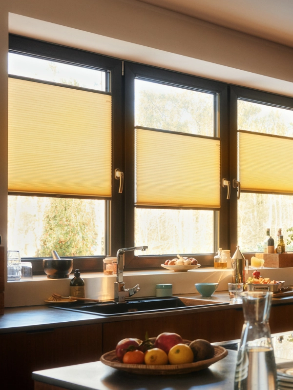 Drei Fenster sind mit gelben Wabenplissees ausgestattet. Sie sind alle eher mittig angeordnet. Direkt angeschlossen ist die Küchenzeile mit Spüle und Kochutensilien. Im Vordergrund ist eine Kücheninsel mit Obstschale und Wasserkaraffe.