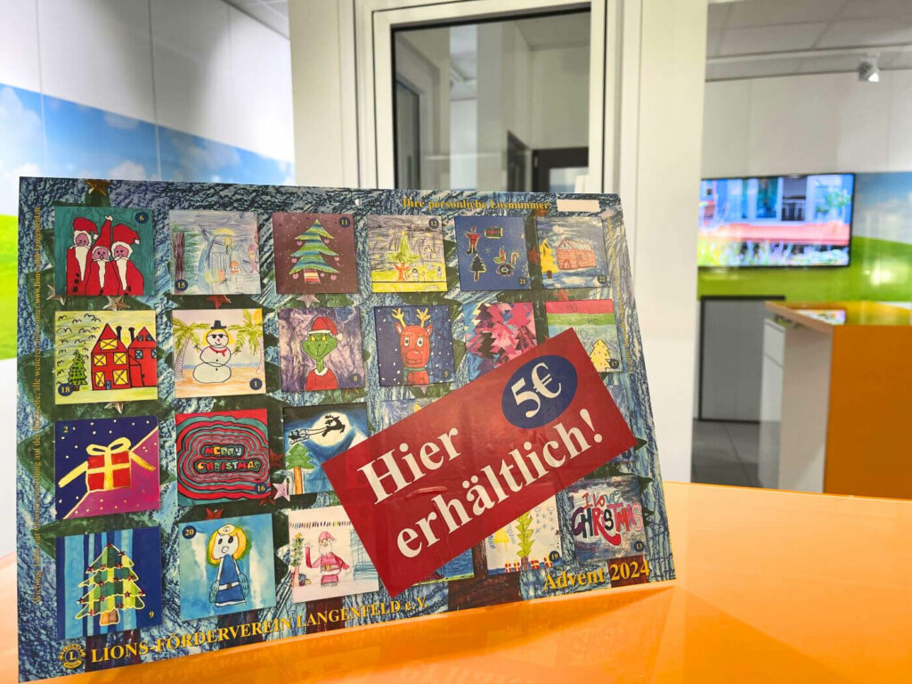 Adventskalender 2024 mit Kinderzeichnungen und rotem Aufkleber „Hier erhältlich! 5 €“ auf orangefarbener Theke im Innenraum.