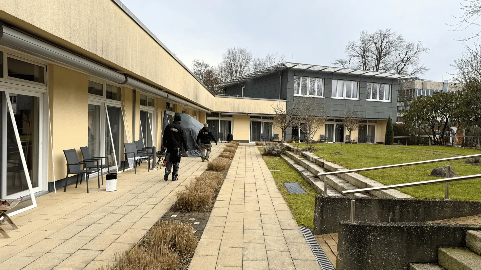 Gebäude der Palliativstation des Klinikums Leverkusen. Links stehen große Spannrahmen an den Fenstern bereit, um eingebaut zu werden. Zwei Monteure der Firma Schulz-Dobrick laufen den Weg entlang.