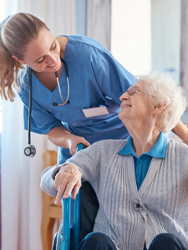 Eine Pflegerin steht hinter einer Seniorin, die im Rollstuhl sitzt. Die Pflegerin trägt einen blauen Kasack und ein Stethoskop um den Hals. Sie beugt sich lächelnd zur Seniorin herunter. Die Seniorin hat weiße, kurze, gelockte Haare und trägt eine Brille. Sie dreht sich lächelnd zur Pflegerin.