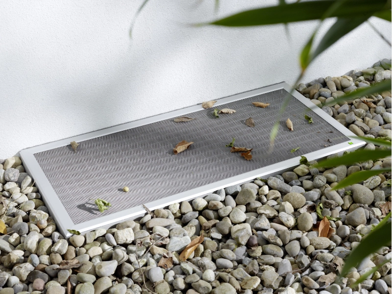 Lichtschachtabdeckung mit stabilem silbernem Gewebe und weißem Rahmen, umgeben von Kieselsteinen und Laubblättern.