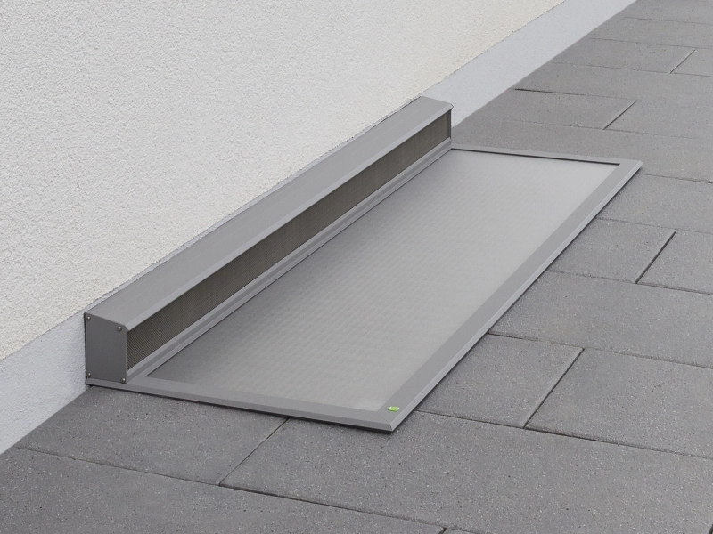 Begehbare Lichtschachtabdeckung aus grauem, robustem Gewebe, bündig installiert auf anthrazitfarbenen Bodenplatten vor einer Wand.