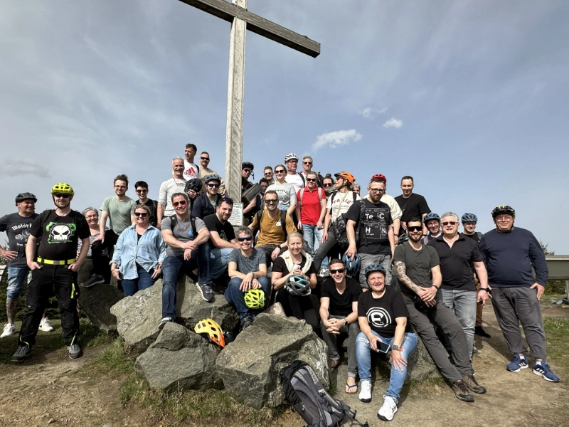 Das Schulz Dobrick Team posiert lächelnd in Fahrradkleidung vor einem großen Kreuz auf einem Felsen während eines gemeinsamen Ausflugs.