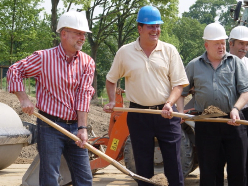 Mehrere Männer in Bauhelmen führen symbolisch den ersten Spatenstich auf einer Baustelle durch.