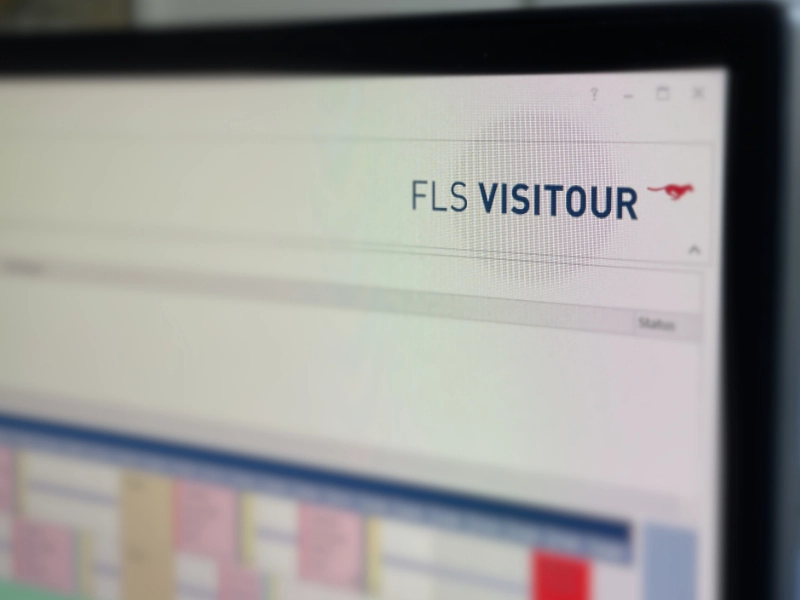 Ein Bildschirm zeigt die Software FLS VISITOUR mit unscharfen Kalender- und Planungsinformationen im Hintergrund.