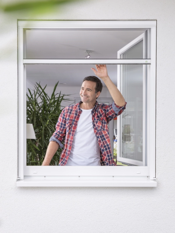 Mann in kariertem Hemd öffnet ein Fliegengitter nach Maß an einem Fenster, umgeben von Zimmerpflanzen.
