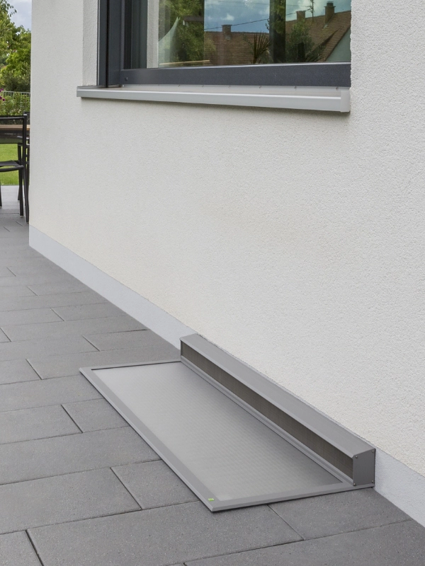 Begehbare Lichtschachtabdeckung in grauer Farbe, bündig in den Boden eingebaut, an einer hellen Hausfassade.