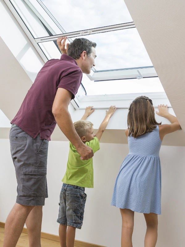 Ein Mann und zwei Kinder öffnen ein Fliegengitter am Dachfenster und schauen gemeinsam nach draußen.