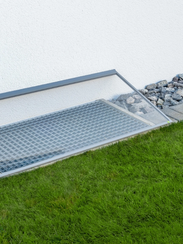 Lichtschachtabdeckung mit Regenschutz aus Glas, sorgt für Belüftung und Schutz vor Regen und Schmutz, eingebaut in einem gepflegten Garten.