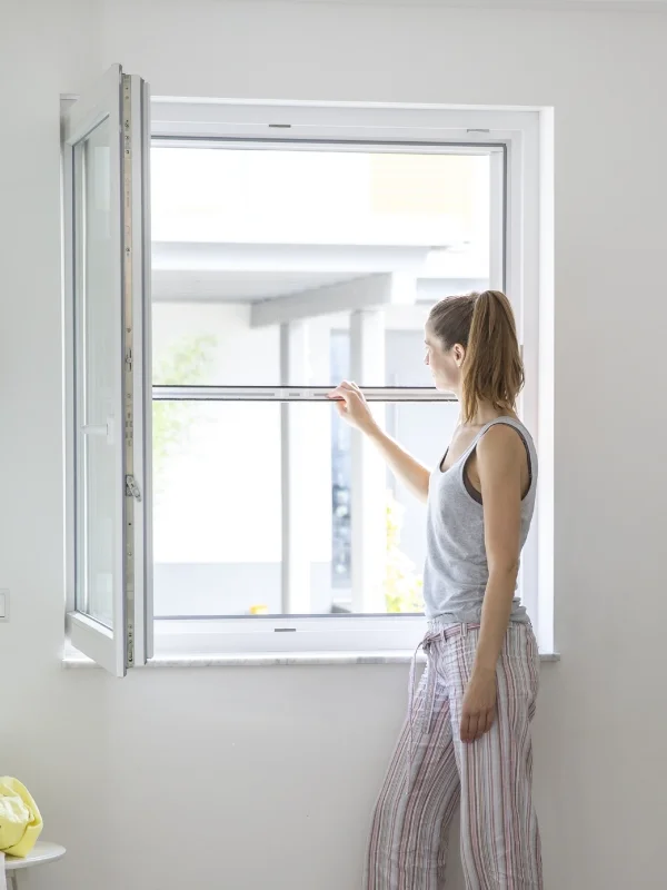 Eine Frau öffnet ein weißes Fenster mit Fliegengitter in einem Raum und blickt nach draußen.