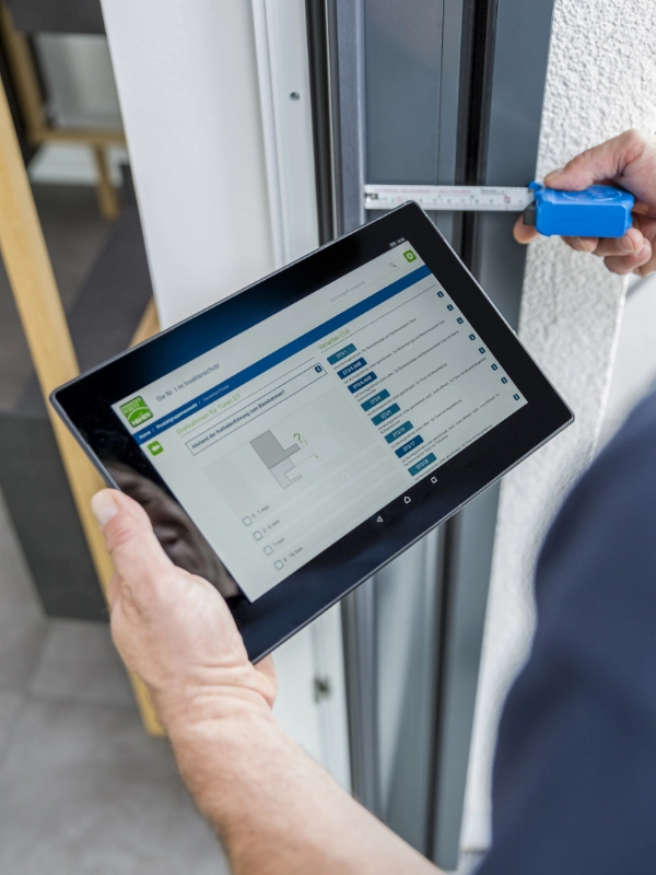 Eine Person misst einen Türrahmen mit einem Maßband und hält dabei ein Tablet mit Planungssoftware von Schulz Dobrick.