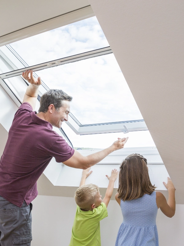 Ein Mann mit zwei Kindern öffnet ein Fliegengitter an einem Dachfenster in einem hellen Raum.