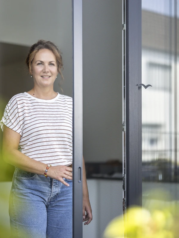 Frau öffnet ein Insektenschutzgitter in einem weißen Fenster und lächelt nach draußen.