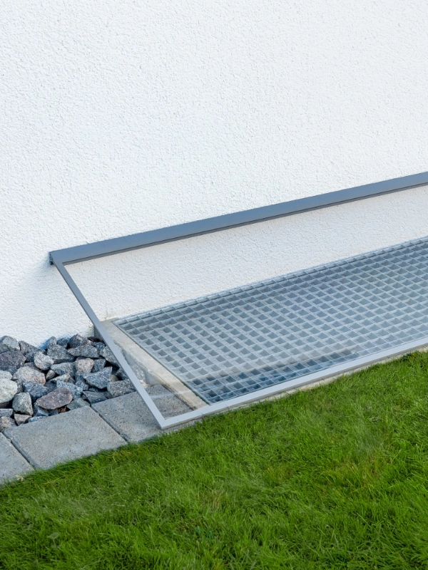 Lichtschachtabdeckung mit Regenschutz neben einer Hauswand, umgeben von Kies und Rasenfläche.