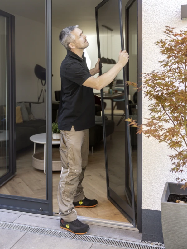 Mann mit Salatschüssel öffnet eine Fliegengitter-Tür nach Maß, während er von einem Garten ins Haus tritt.
