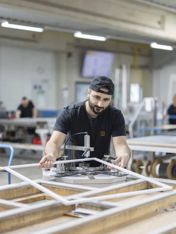 Mitarbeiter arbeiten in einer Produktionshalle an Fliegengittern, während ein Arbeiter im Vordergrund ein Gestell transportiert.