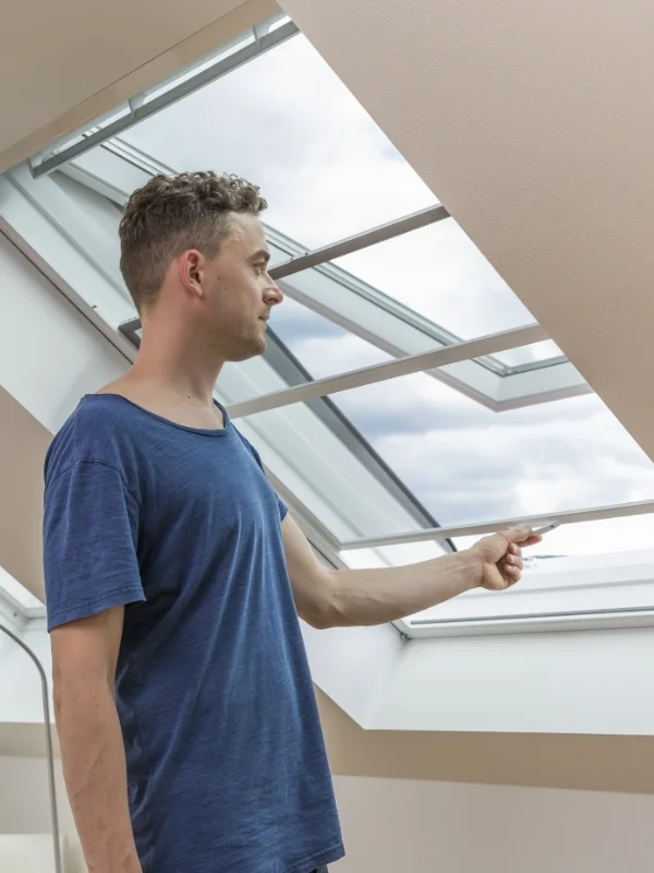 Ein Mann in einem blauen T-Shirt öffnet ein Fliegengitter am Dachfenster in einem Raum.
